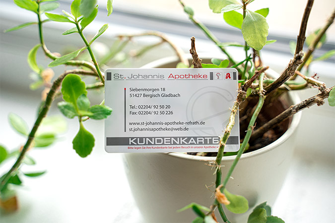 Kundenkarte der St. Johannis Apotheke in Bergisch Gladbach, eingebettet im Gestüpp einer Pflanze